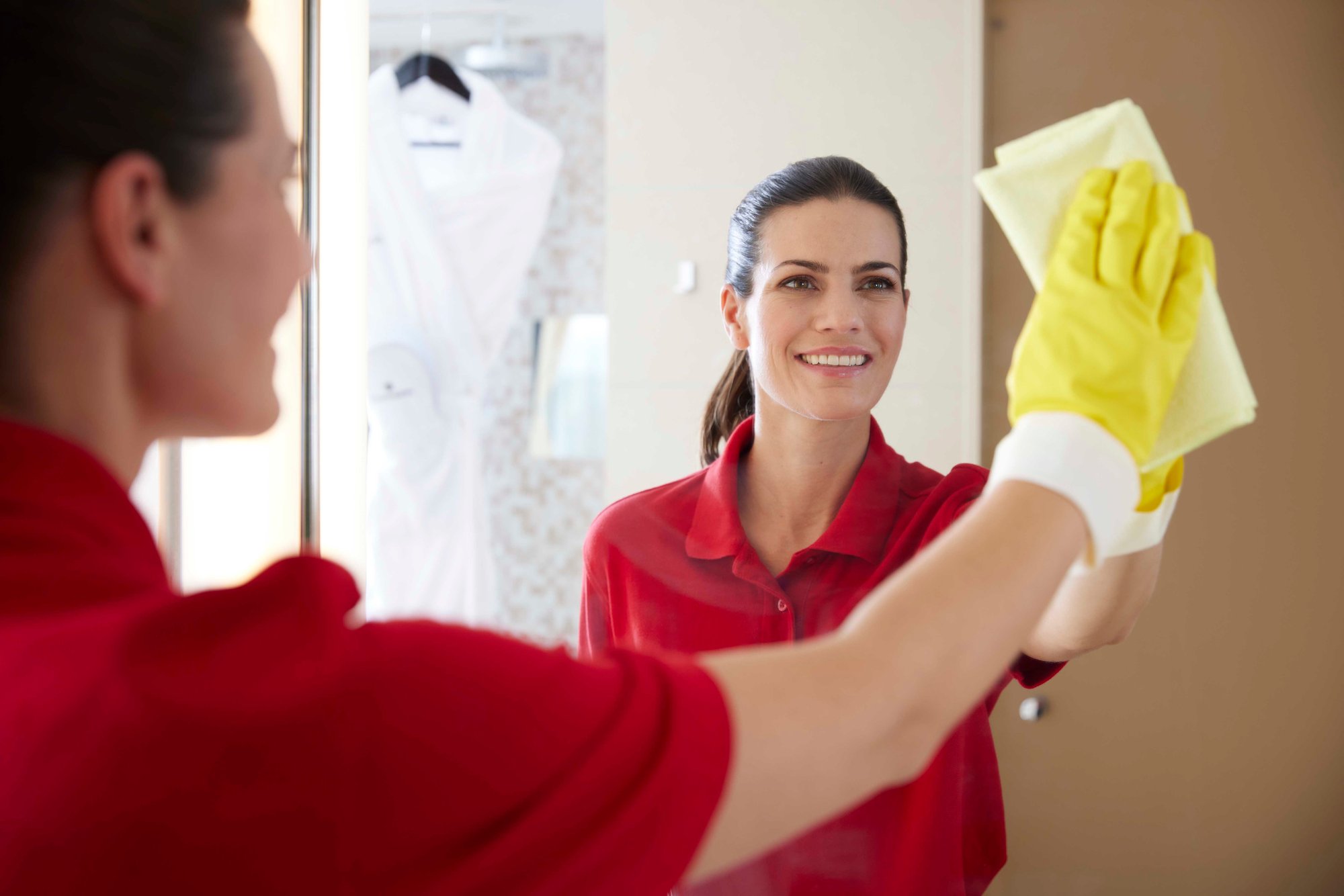 A housekeeper wearing a red polo shirt and yellow Vileda Professional gloves smiles while cleaning a mirror with a yellow microfiber cloth. The reflection shows her focused expression and the clean, modern bathroom environment, including a white bathrobe hanging in the background.